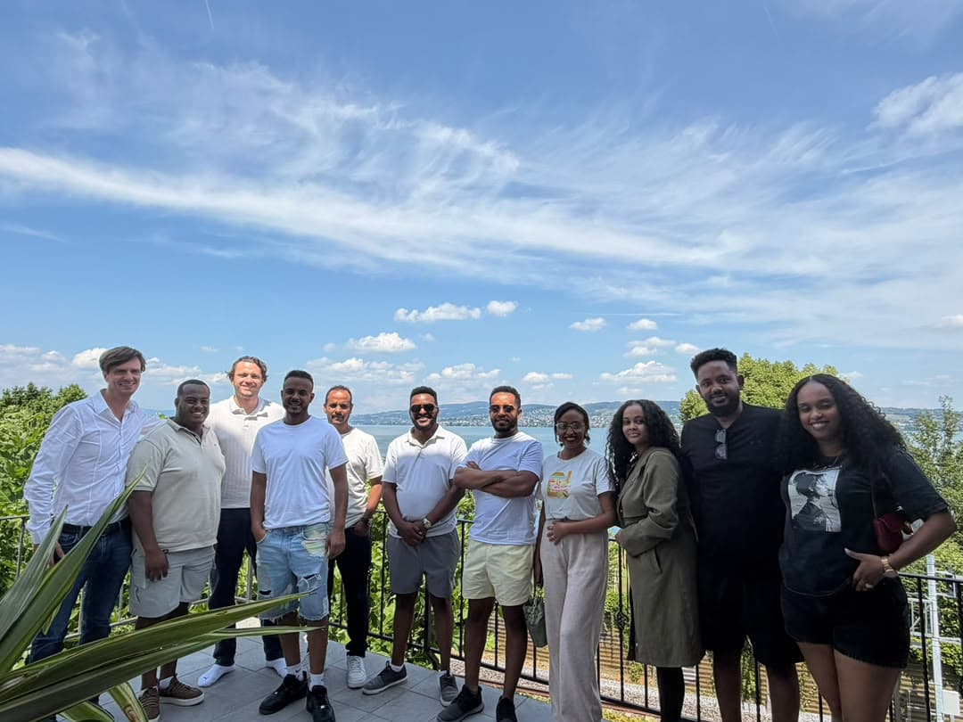 A group of people smiling together on a terrace, with a lake and a bright blue sky behind them